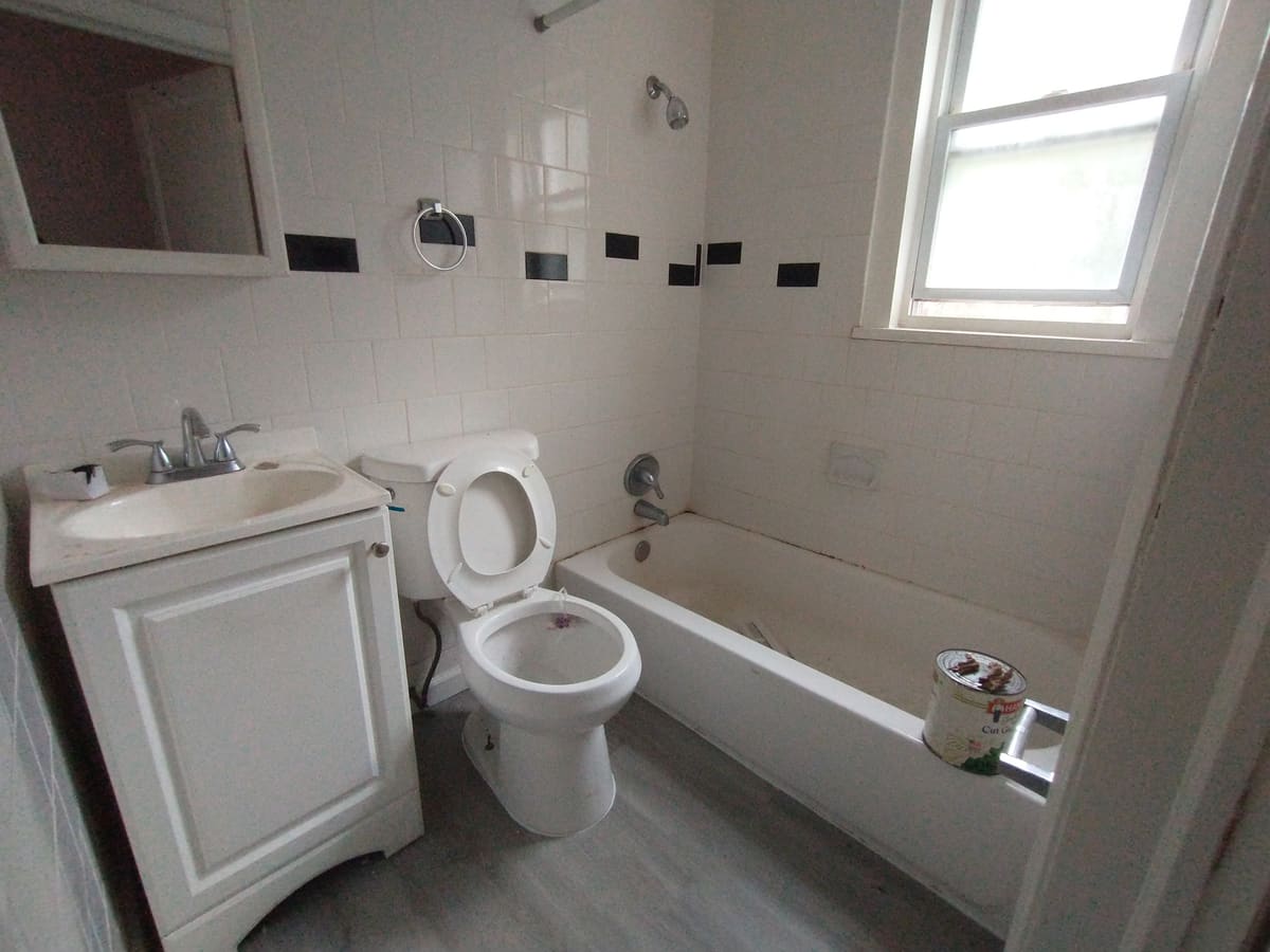 Original unit bathroom before renovation: dated white vanity, black accent tile border, worn tub surround and vinyl flooring.