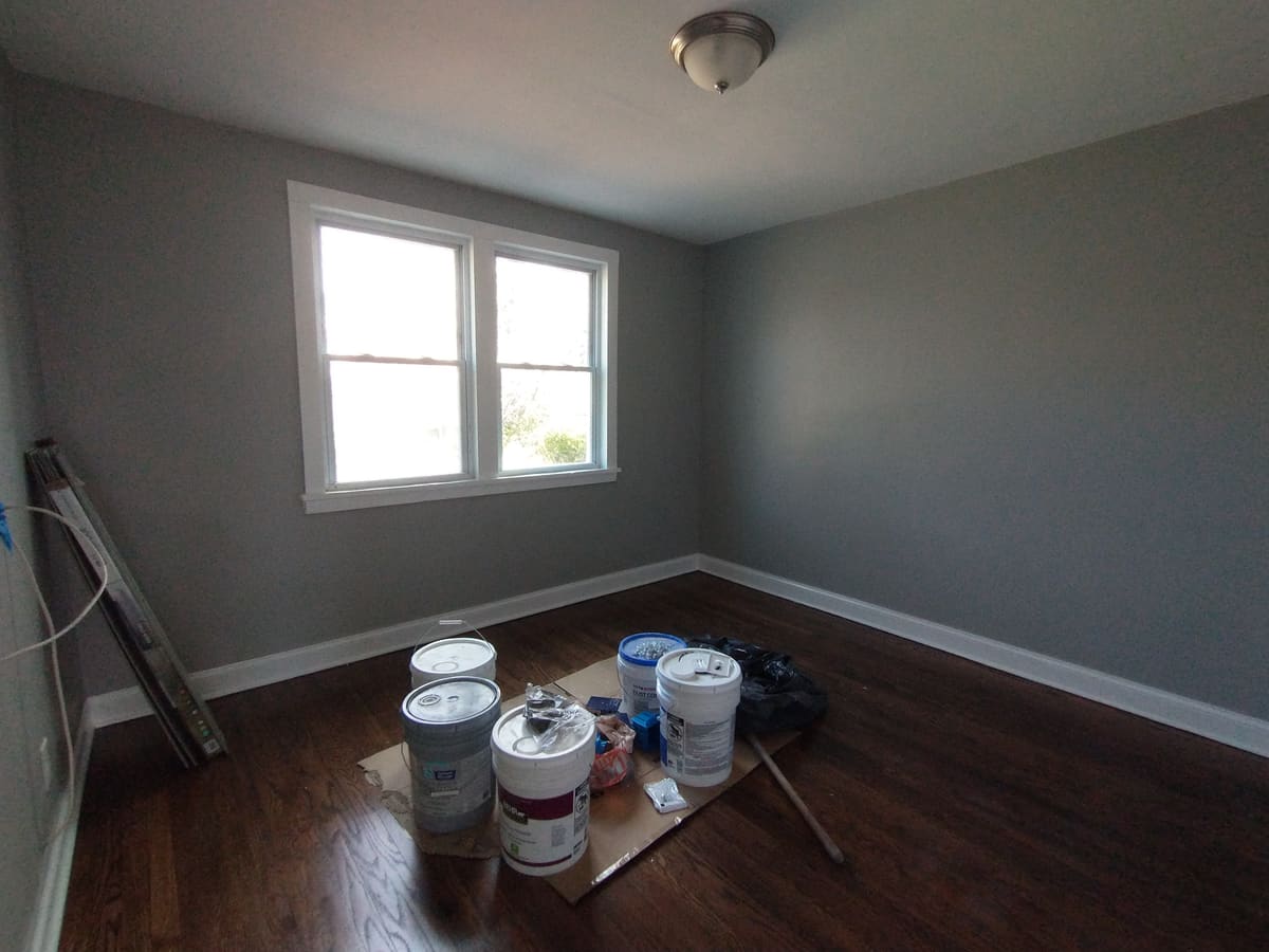 Bedroom under renovation with refinished dark hardwood, freshly painted grey walls, and paint buckets and materials staged on the floor.