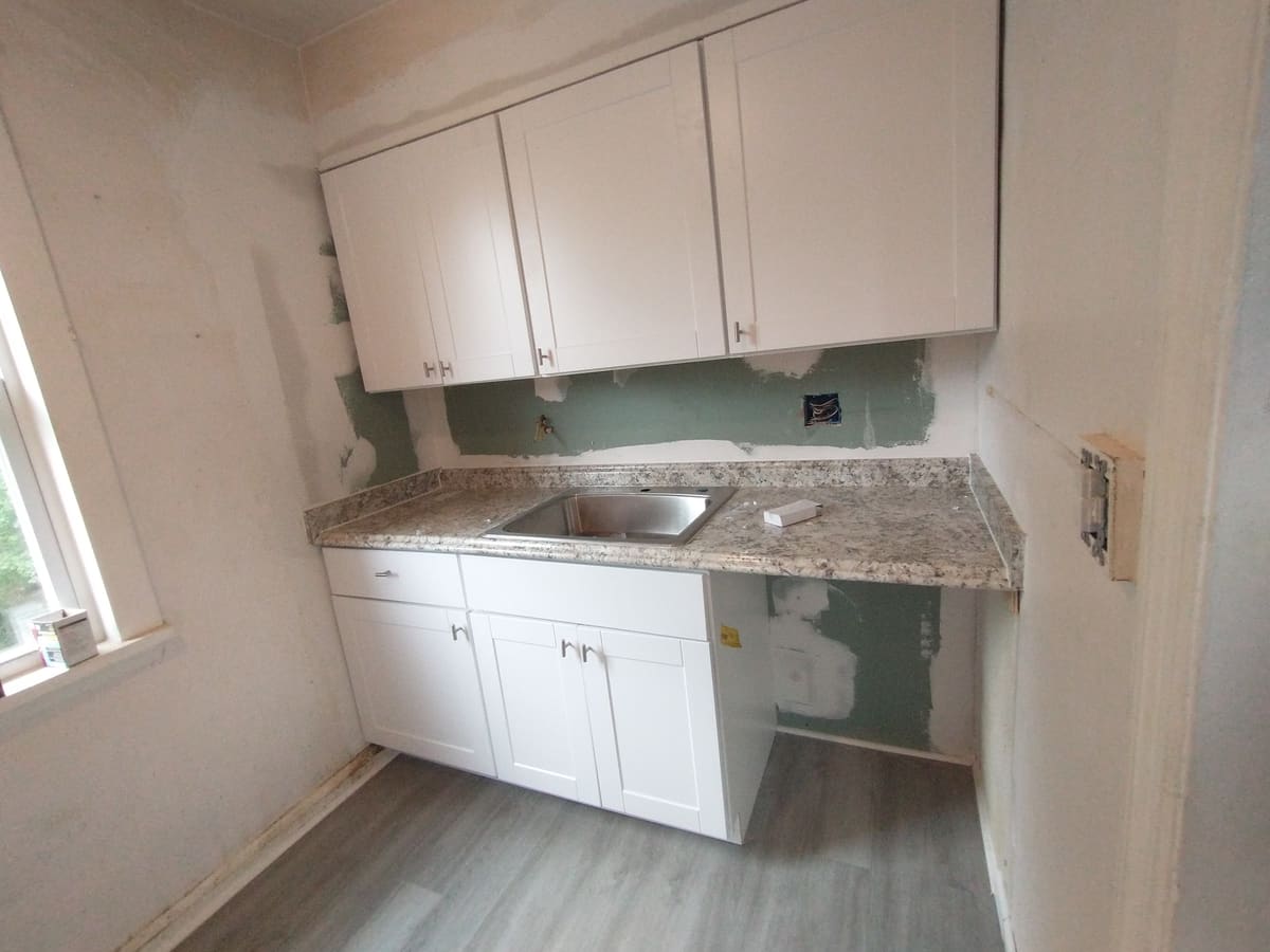 New white shaker cabinets and granite countertop installed over unfinished patched drywall, with new plank flooring down and the sink not yet plumbed.